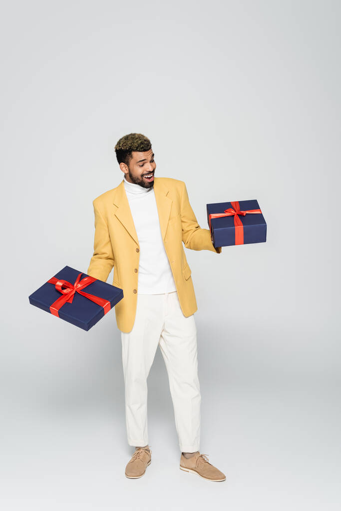 pleine longueur de l'homme afro-américain excité en jaune veston élégant tenant des cadeaux enveloppés sur gris  - Photo, image