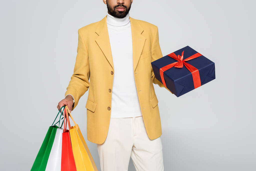 Sarı ceketli, sakallı Afro-Amerikan bir adamın hediyesini ve gri üzerinde izole edilmiş alışveriş çantalarını...  - Fotoğraf, Görsel