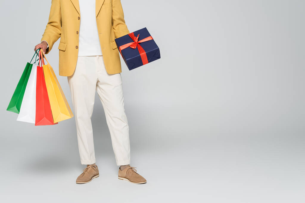 vista cortada do homem americano africano em blazer elegante amarelo segurando presente embrulhado e sacos de compras isolados em cinza  - Foto, Imagem