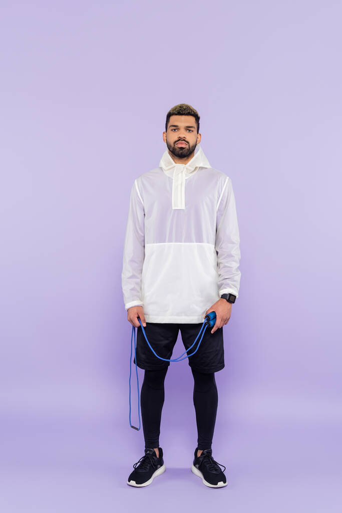 longitud completa de joven afroamericano hombre en ropa deportiva de moda celebración de saltar la cuerda en púrpura - Foto, Imagen