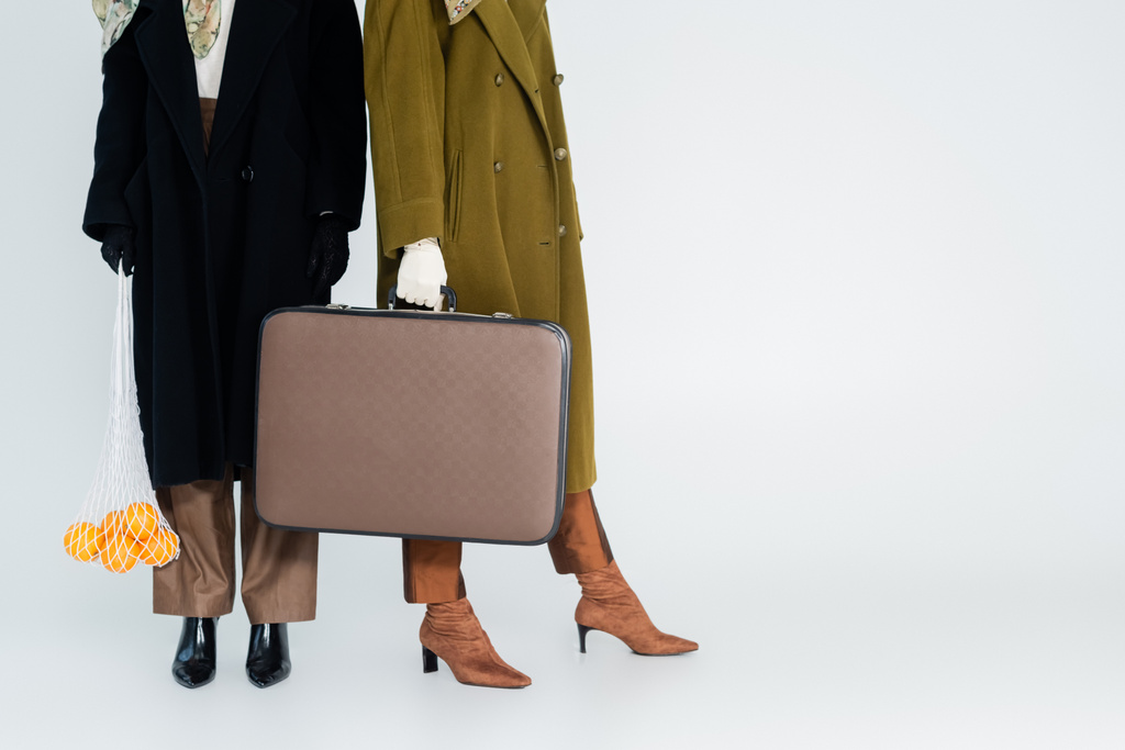 グレーの背景にオレンジとヴィンテージスーツケースとネットバッグを保持コートの女性の部分的なビュー - 写真・画像