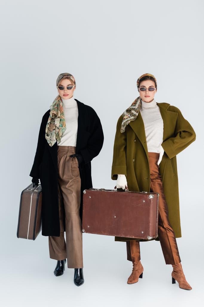 teljes hossza stílusos nők kabát és napszemüveg pózol vintage bőröndök szürke alapon - Fotó, kép