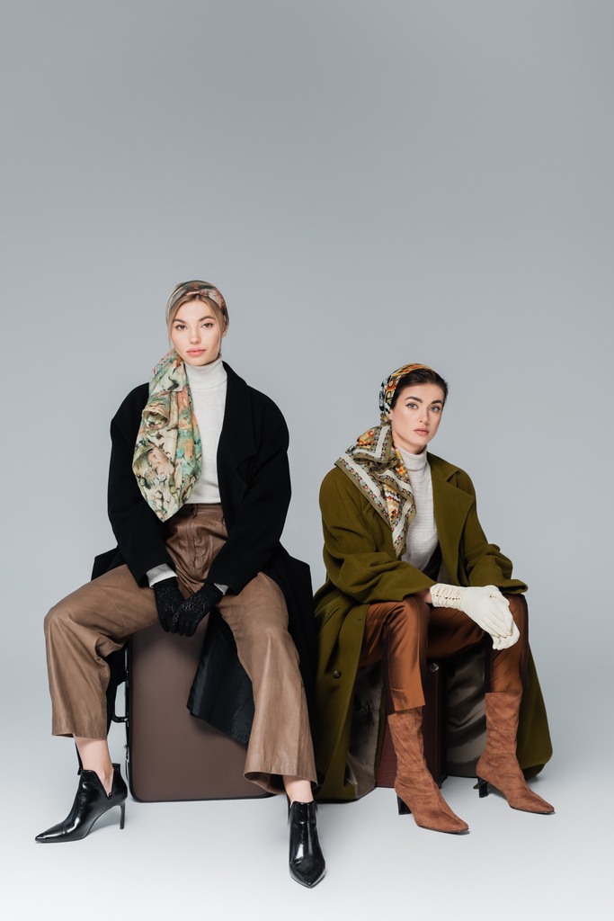 longitud completa de las mujeres de moda sentadas en maletas vintage sobre fondo gris - Foto, Imagen