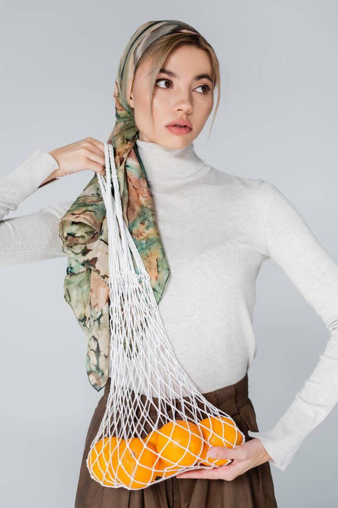 žena ve stylovém šátku pózující s čerstvými pomeranči v síťované tašce izolované na šedé - Fotografie, Obrázek