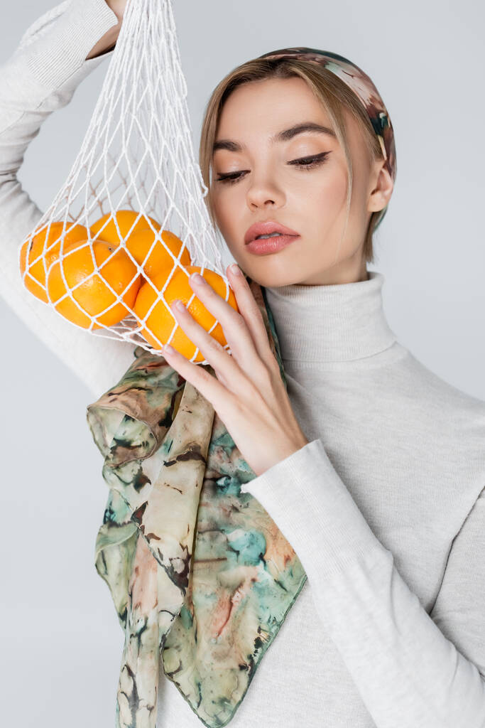 красивая женщина в платке позирует со свежими апельсинами в сетчатом мешке, изолированном на сером - Фото, изображение