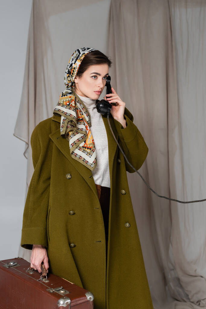 mujer con abrigo verde y pañuelo estampado sosteniendo la maleta y hablando por teléfono vintage sobre fondo gris drapeado - Foto, Imagen