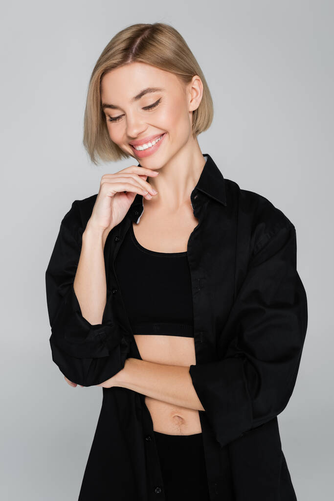 hübsche Frau in schwarzem BH und Hemd, die das Kinn berührt und isoliert auf grau lächelt - Foto, Bild