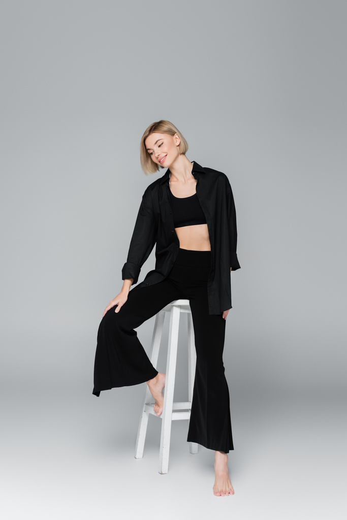 full length of smiling woman in black trendy clothes posing on white stool on grey background - Photo, Image