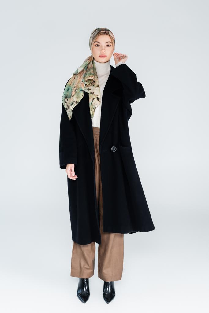 άποψη πλήρους μήκους της γυναίκας με μαντίλα και μαύρο παλτό στέκεται σε γκρι φόντο - Φωτογραφία, εικόνα