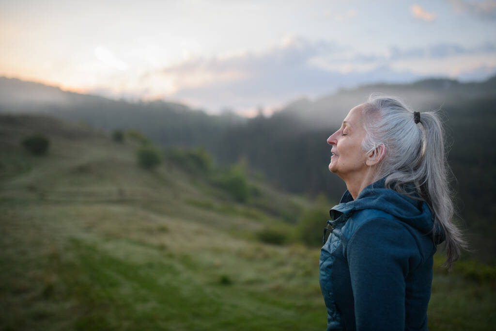 Vanhempi nainen tekee hengitys liikuntaa luonnossa varhain aamulla sumu ja vuoret taustalla. - Valokuva, kuva
