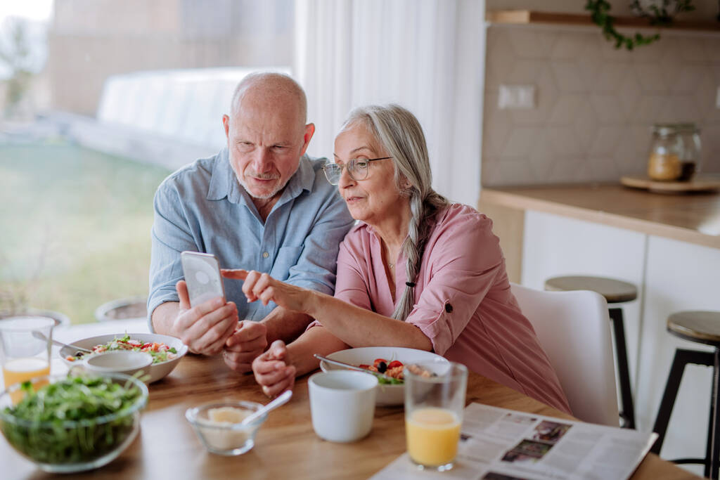 Una feliz pareja de personas mayores usando un smartphone durante la cena juntos en casa. - Foto, Imagen