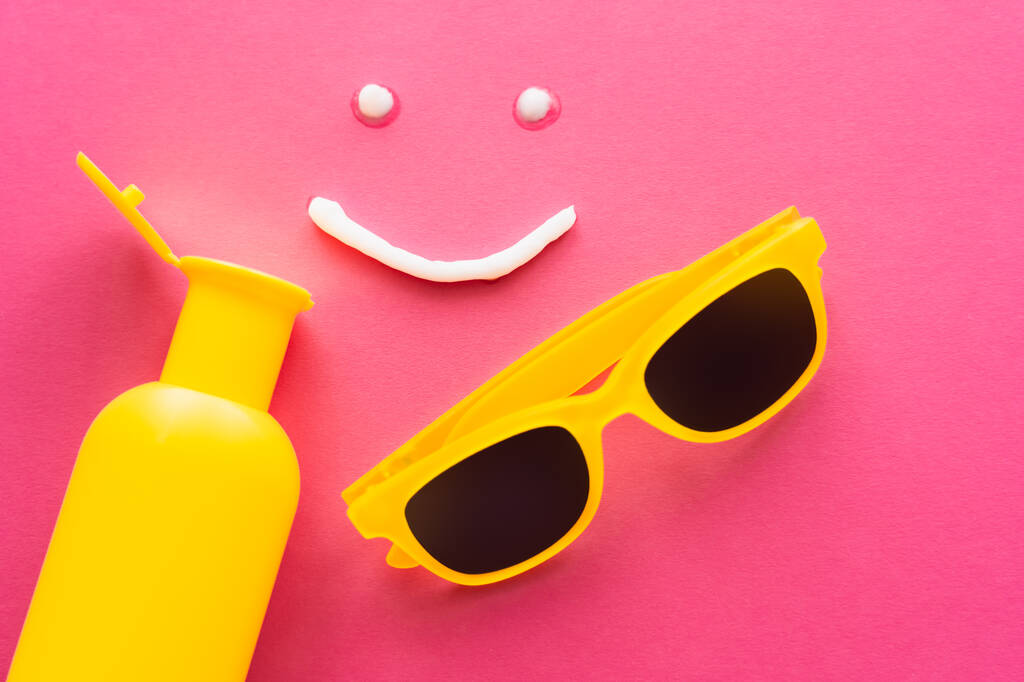 Vista superior del signo de sonrisa del protector solar cerca de la botella y gafas de sol sobre fondo rosa  - Foto, imagen