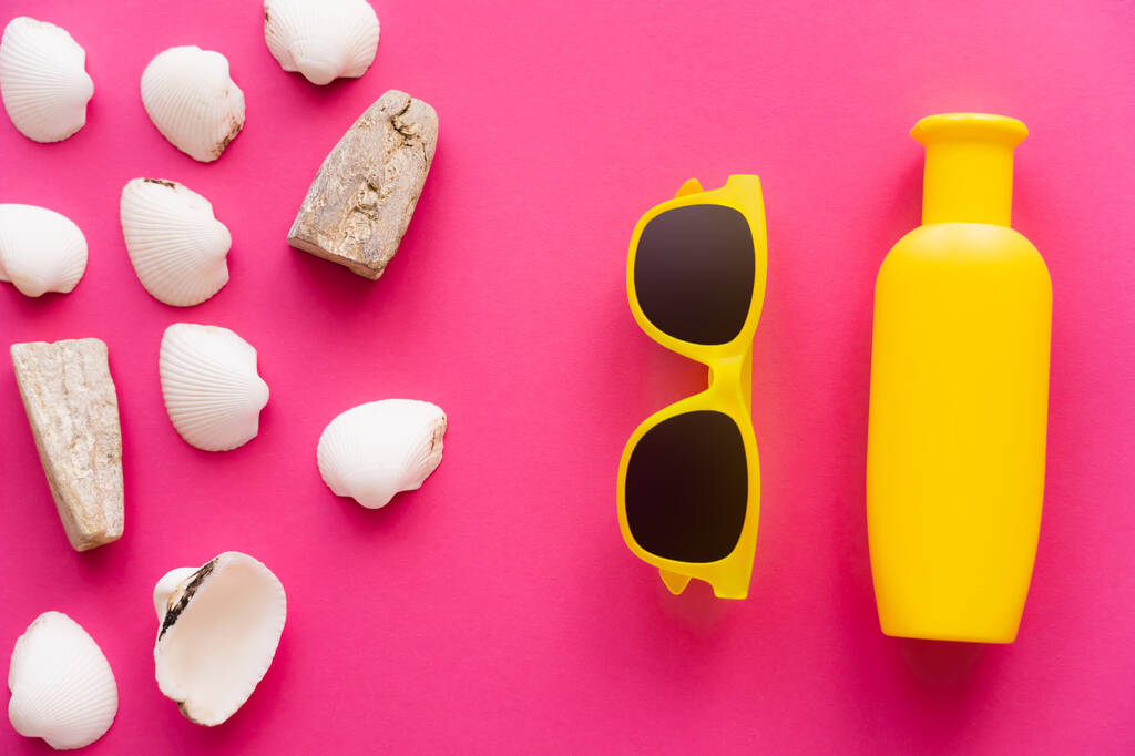Vista superior de conchas perto de óculos de sol e garrafa de protetor solar em fundo rosa  - Foto, Imagem