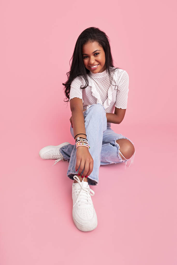 Jovencita alegre en jeans y blusa con zapatillas blancas sonriendo y mirando a la cámara mientras está sentada sobre fondo rosa - Foto, imagen