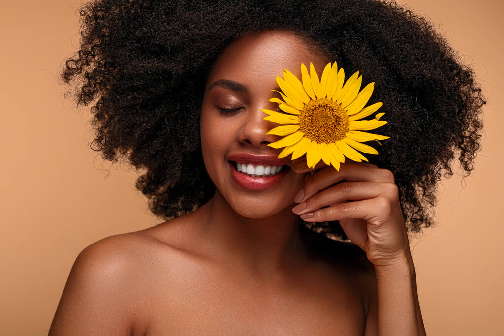 Vrolijke jonge zwarte vrouw met blote schouders en Afro kapsel glimlachend en bedekt gesloten ogen, met frisse zonnebloem tijdens huidverzorging routine tegen bruine achtergrond. - Foto, afbeelding