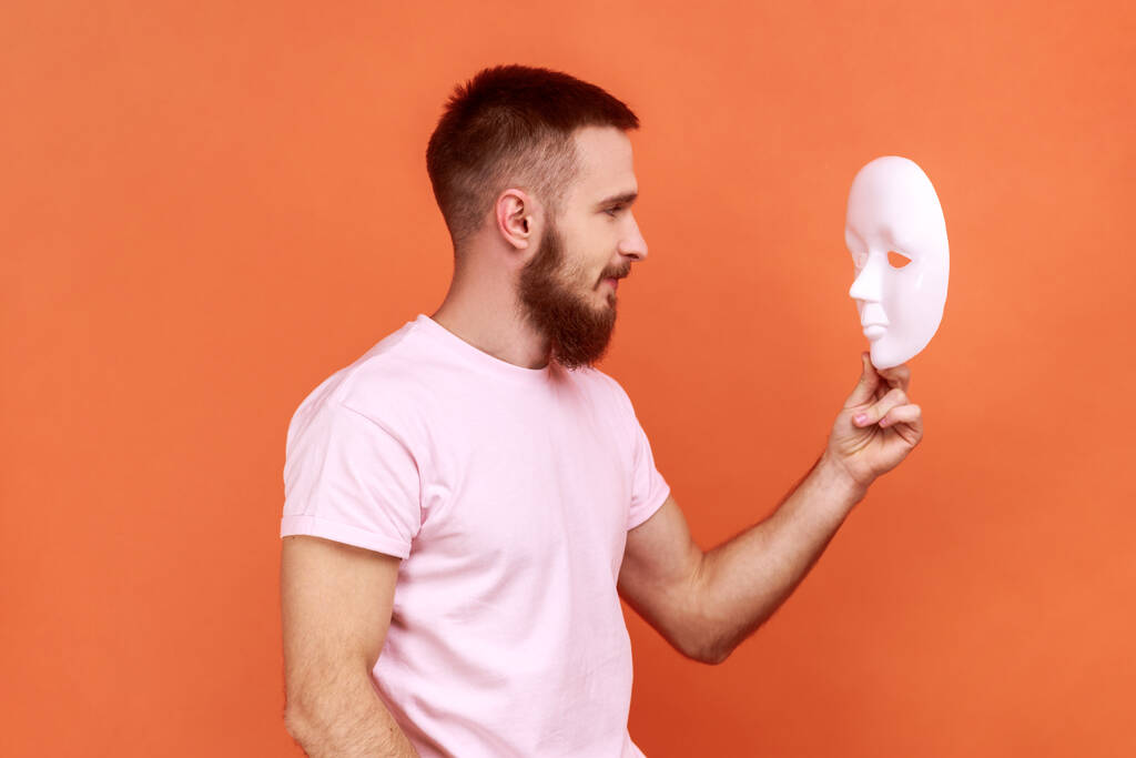Boční pohled na portrét vousatého muže s pozorným výrazem hledícího na bílou masku v rukou, snažícího se pochopit skrytou osobnost, oblečeného v růžovém tričku. Vnitřní studio záběr izolované na oranžovém pozadí. - Fotografie, Obrázek