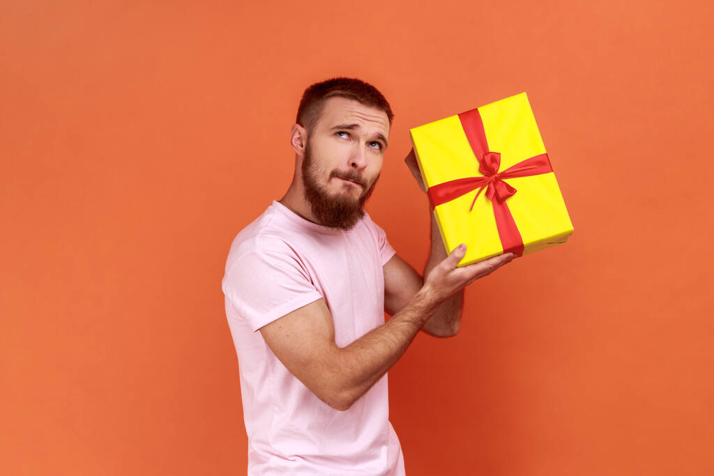 Portrait of curious bearded man standing, holding yellow gift box, looking away with pensive expression, thinks what inside, wearing pink T-shirt. Indoor studio shot isolated on orange background. - Photo, Image