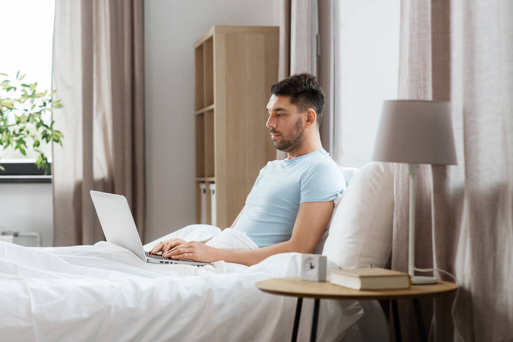 people, technology and remote job concept - man with laptop computer lying in bed at home bedroom - Photo, Image