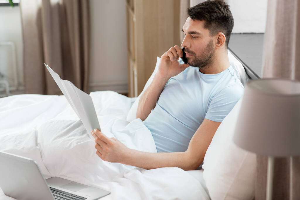 люди, технологии и концепция удаленной работы - человек с папкой и ноутбуком, звонящий по смартфону в постели в домашней спальне - Фото, изображение