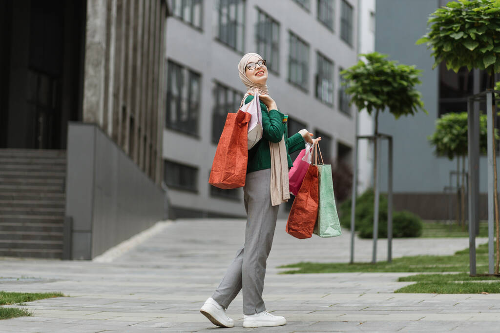 Jolie jeune femme musulmane, vêtue d'un pantalon gris et d'une veste verte, marchant dans la rue avec des sacs colorés, souriant et regardant dehors. Shopping dans le concept de la ville. - Photo, image