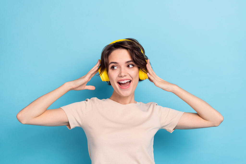 Фото блискучої збудженої жінки, що носить бежеві футболки навушники, насолоджуючись музикою, яка виглядає порожнім простором ізольованим синім кольором фону
. - Фото, зображення