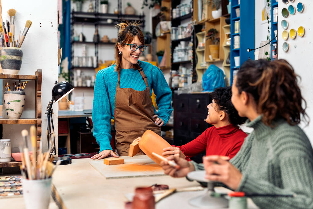 Photo de stock de femmes heureuses dans le tablier en utilisant de l'argile et de parler pendant le cours de poterie. - Photo, image