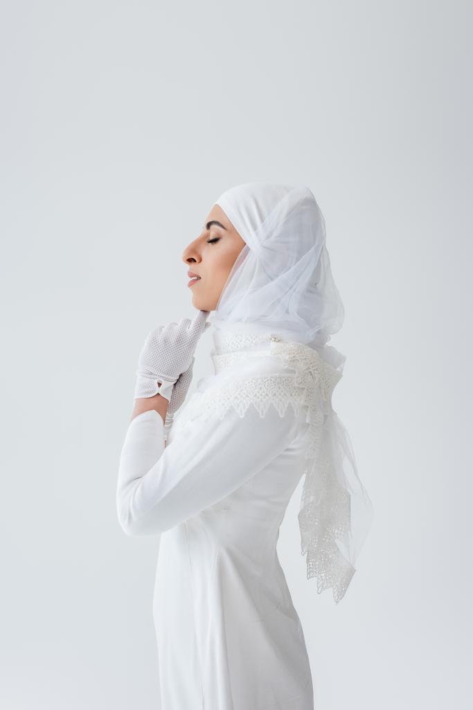 вид сбоку мусульманской невесты в перчатках и свадебном платье, молящейся изолированно на сером - Фото, изображение