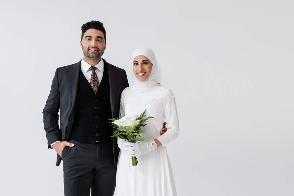 muzułmańska panna młoda w rękawiczkach i sukni ślubnej z bukietem calla lilia w pobliżu szczęśliwego pana młodego w garniturze odizolowanym na szaro - Zdjęcie, obraz