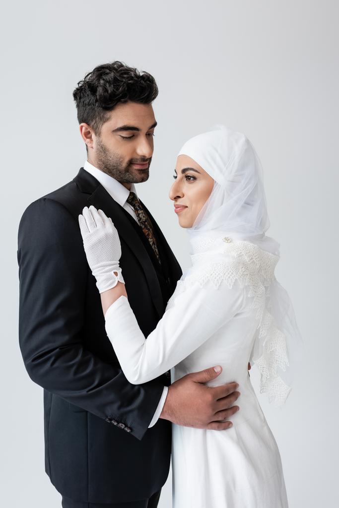 młody muzułmanin panna młoda w hidżab i suknia ślubna i pan młody przytulanie izolowany na szary - Zdjęcie, obraz