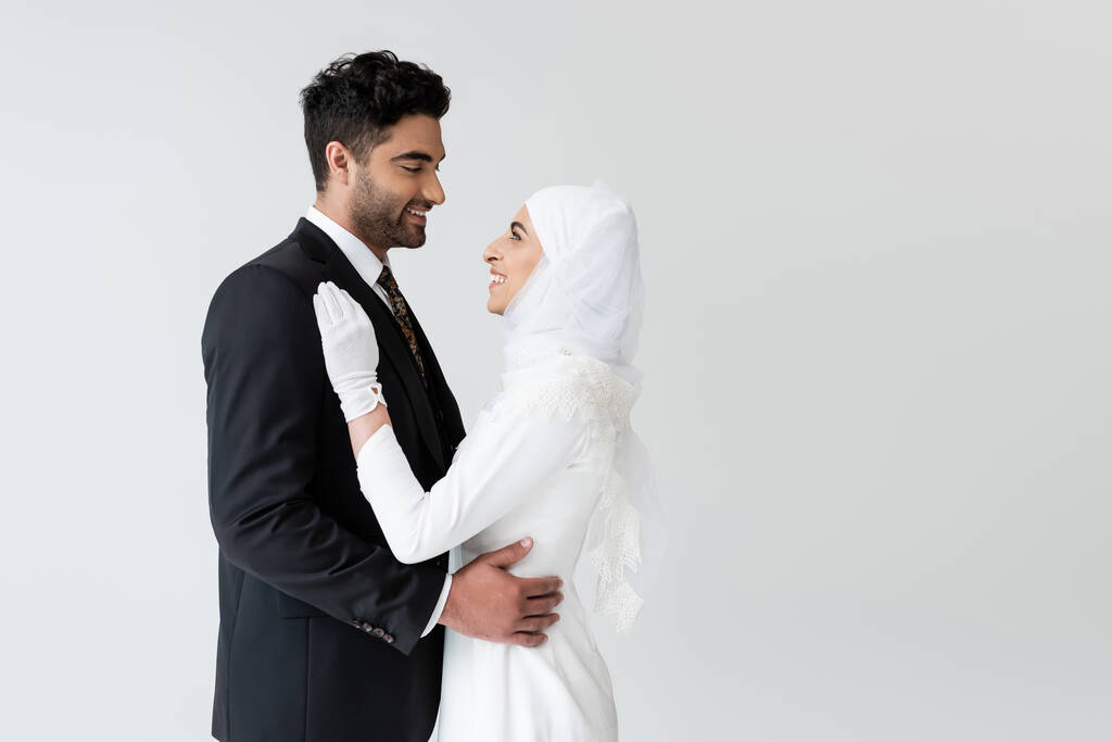 glückliche muslimische Braut in Hijab und Hochzeitskleid und Bräutigam umarmen sich, während sie sich isoliert auf grau anschauen - Foto, Bild