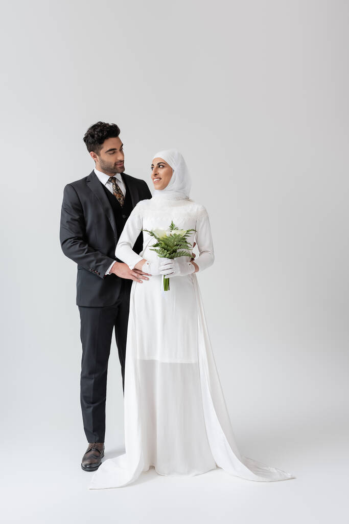 μουσουλμάνος γαμπρός αγκαλιάζει χαρούμενη νύφη σε νυφικό με μπουκέτο σε γκρι  - Φωτογραφία, εικόνα