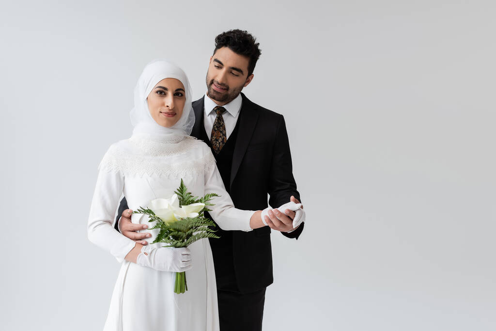 novio musulmán cogido de la mano de la novia sonriente en vestido de novia con ramo aislado en gris  - Foto, Imagen