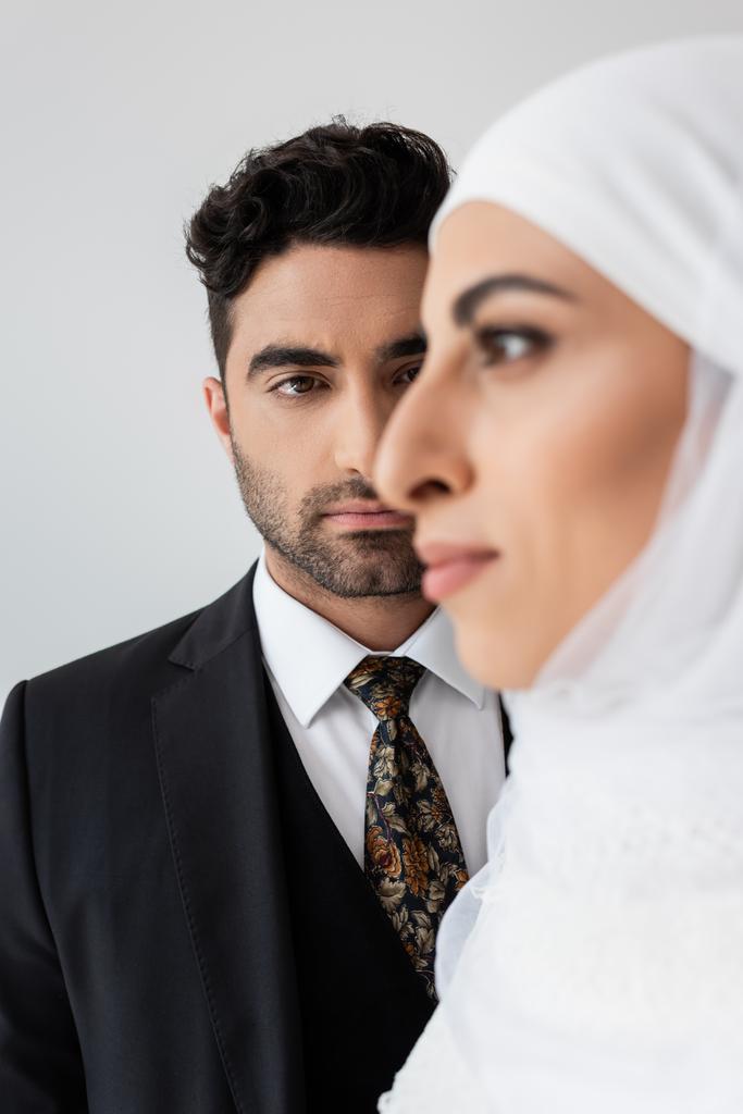 Moslim man kijkt naar wazige bruid op de voorgrond geïsoleerd op grijs - Foto, afbeelding