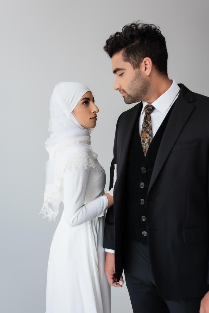 kobieta w sukni ślubnej i hidżab stojący obok męża w garniturze odizolowany na szarości  - Zdjęcie, obraz