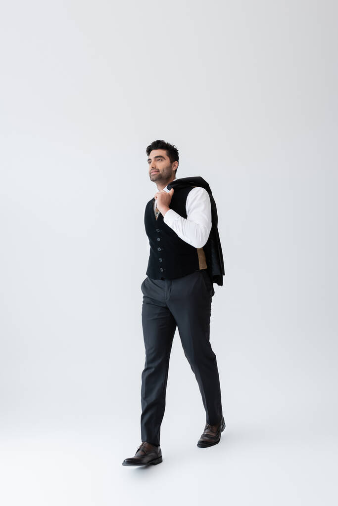 full length of muslim man in vest walking while holding blazer on grey - Photo, Image