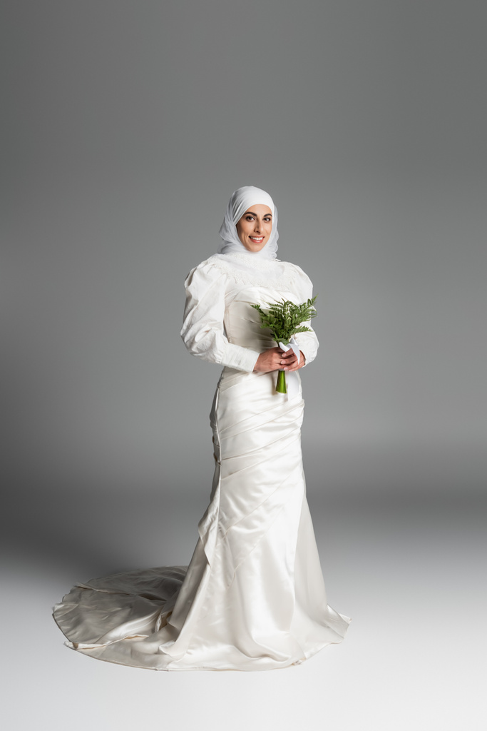 full length of pleased muslim bride σε λευκό φόρεμα και χιτζάμπ κρατώντας γαμήλιο μπουκέτο σε σκούρο γκρι  - Φωτογραφία, εικόνα