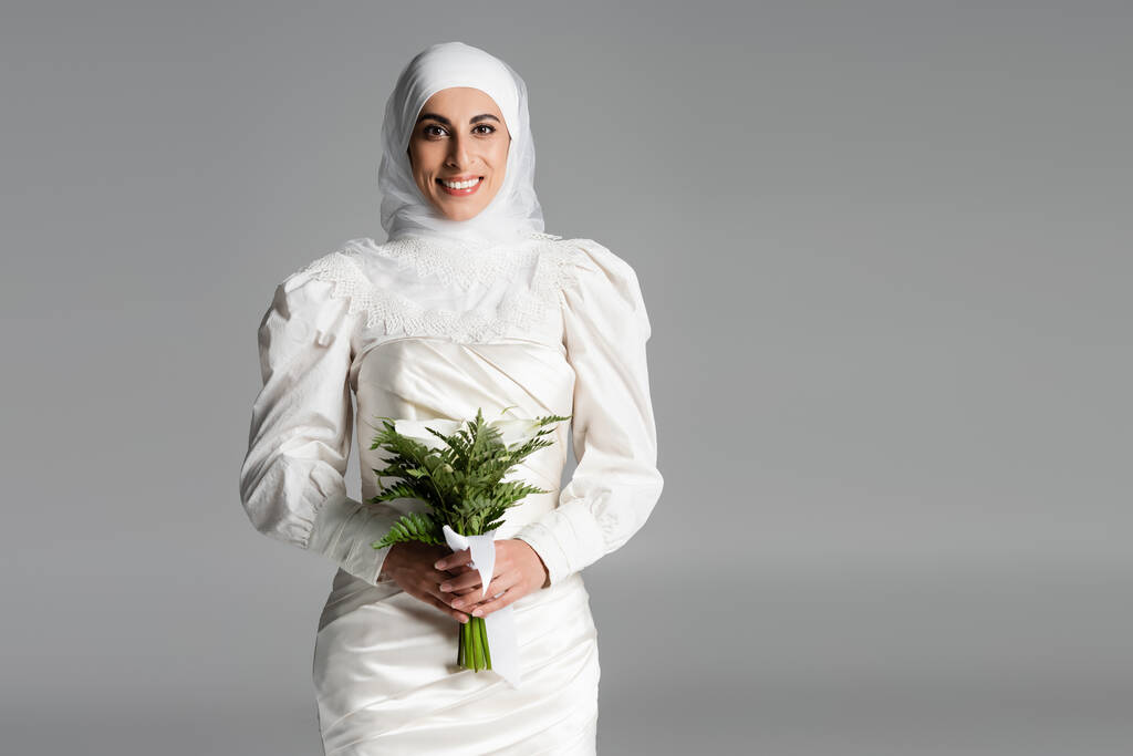 vreugdevolle moslim bruid in witte jurk en hijab houden bruiloft boeket op donkergrijs  - Foto, afbeelding