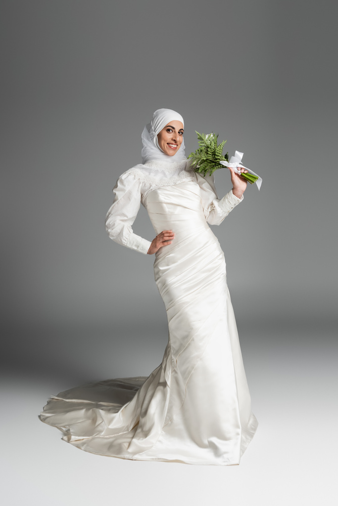 volledige lengte van gelukkig moslim bruid in witte jurk en hijab houden bruiloft boeket terwijl poseren op donkergrijs  - Foto, afbeelding