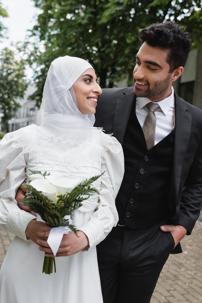 šťastný ženich pózuje s rukou v kapse a při pohledu na muslimskou nevěstu v hidžábu se svatební kyticí  - Fotografie, Obrázek