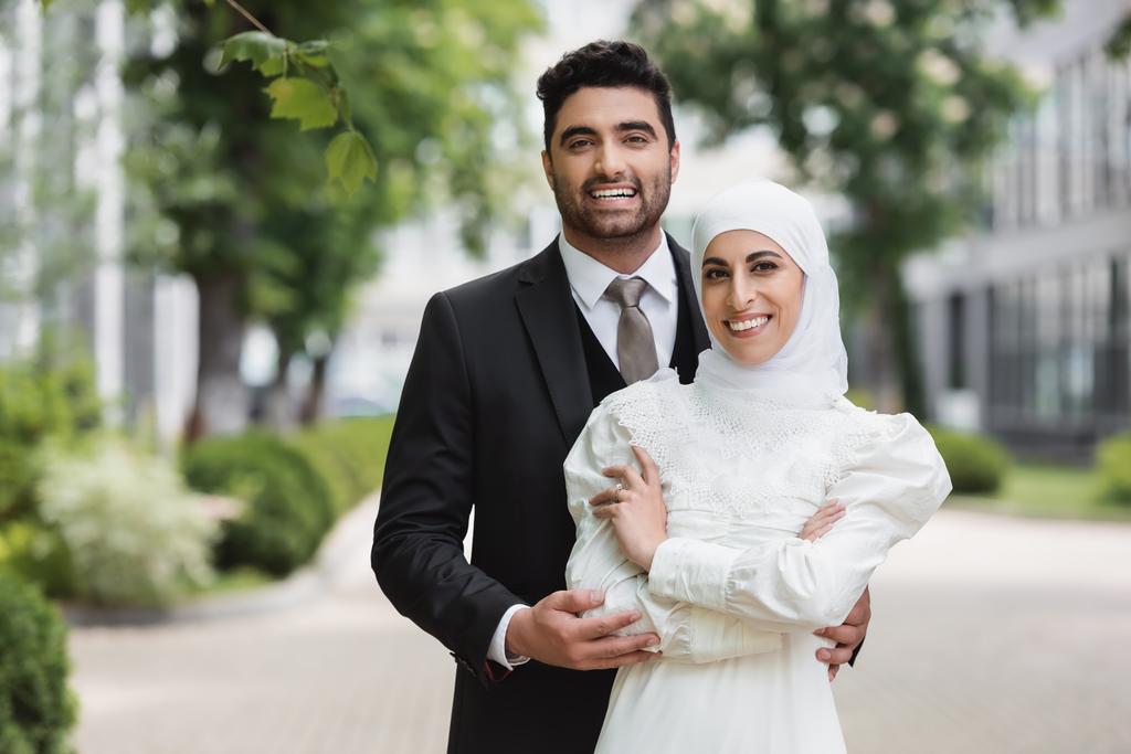 χαρούμενος γαμπρός αγκαλιάζει μουσουλμανική νύφη σε μαντίλα με βέρα  - Φωτογραφία, εικόνα