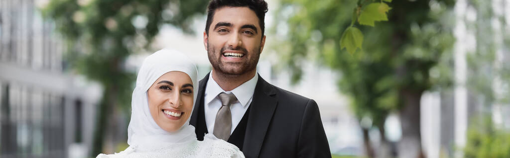 щасливий наречений обіймає мусульманську наречену в хіджабі посміхаючись і дивлячись на камеру, банер
 - Фото, зображення