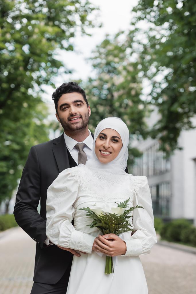 noiva muçulmana alegre no casamento hijab e vestido branco segurando buquê perto do noivo fora  - Foto, Imagem