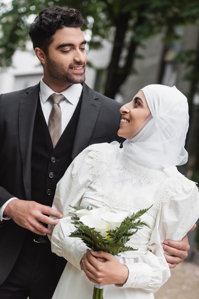 vidám muszlim menyasszony esküvő hidzsáb és fehér ruha gazdaság csokor és nézi a vőlegény kívül  - Fotó, kép