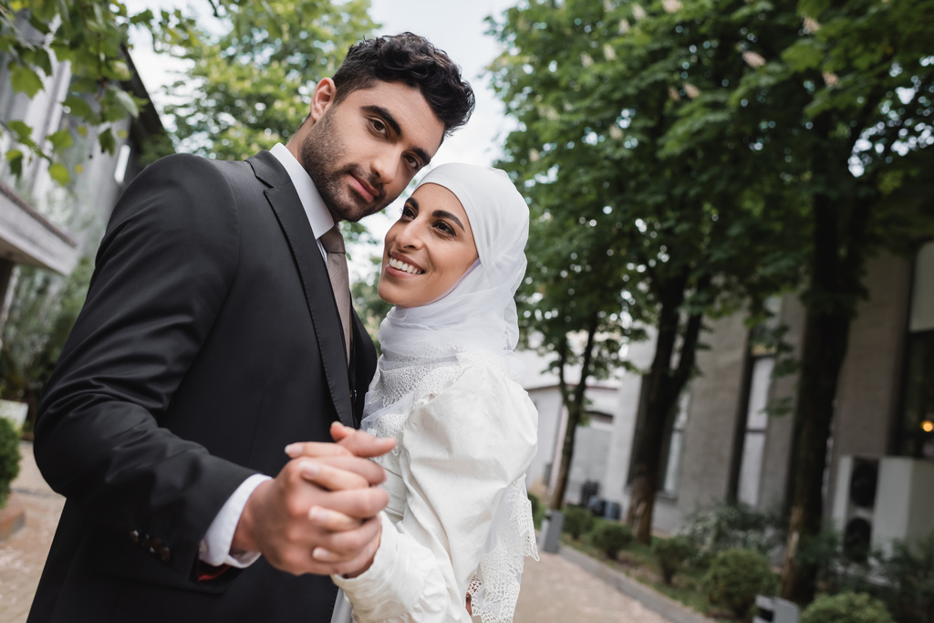 szczęśliwy muzułmanin nowożeńcy trzymając się za ręce i uśmiechając się w zielonym parku  - Zdjęcie, obraz