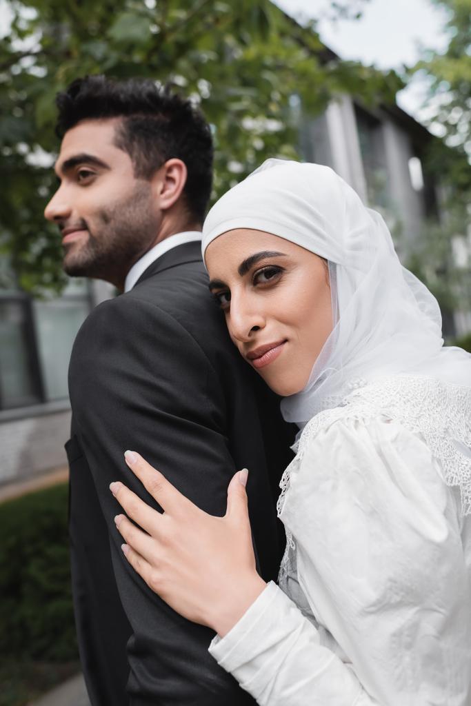 vrolijk moslim bruid in hijab en trouwjurk knuffelen bruidegom  - Foto, afbeelding