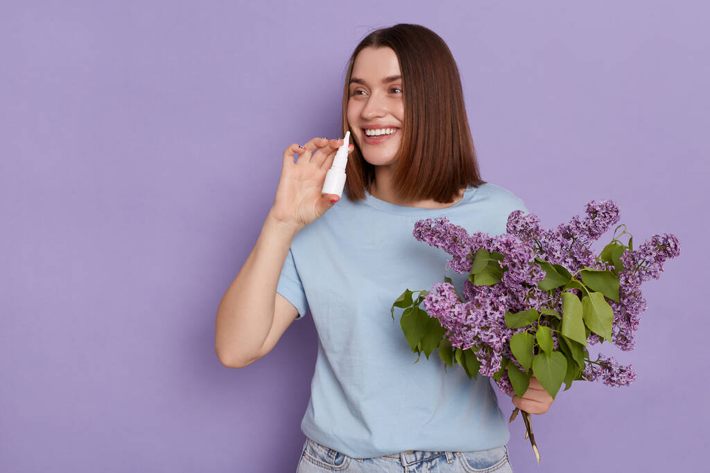 Indoor shot of happy dark haired woman holding nasal spray found proper medicament for stuff nose suffers from allergic rhinitis, posing with lilac bouquet isolated over lilac background. - Photo, Image