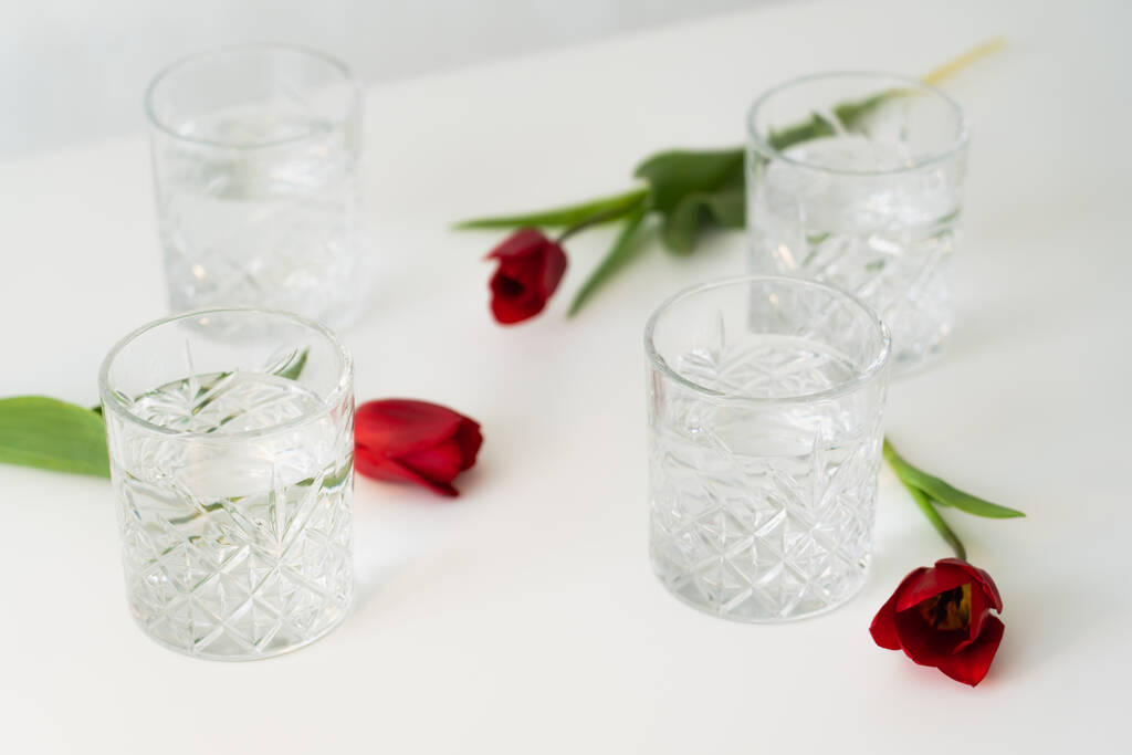 граненые очки с водой рядом с красными тюльпанами на белом размытом фоне - Фото, изображение