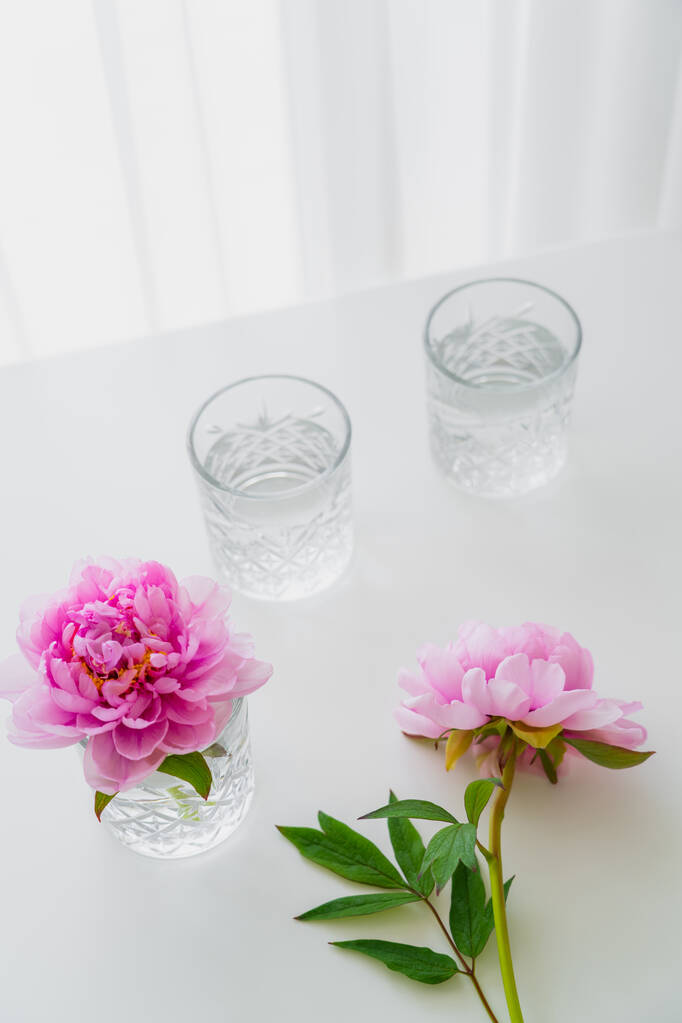 Hochwinkelaufnahme facettierter Gläser mit Wasser und rosa Pfingstrosen auf weißer Oberfläche - Foto, Bild