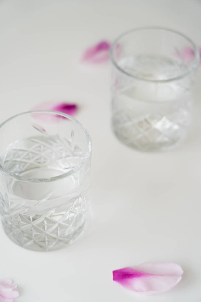 закрыть обзор стаканов с пресной водой рядом с лепестками цветов на белой поверхности и размытым фоном - Фото, изображение
