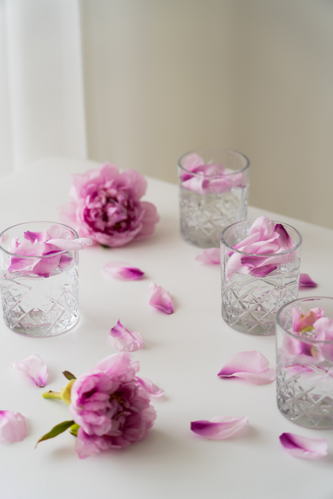 白いテーブルの上にピンクの牡丹の近くに強壮剤と花の花びらを持つ結晶ガラスとグレーの背景 - 写真・画像